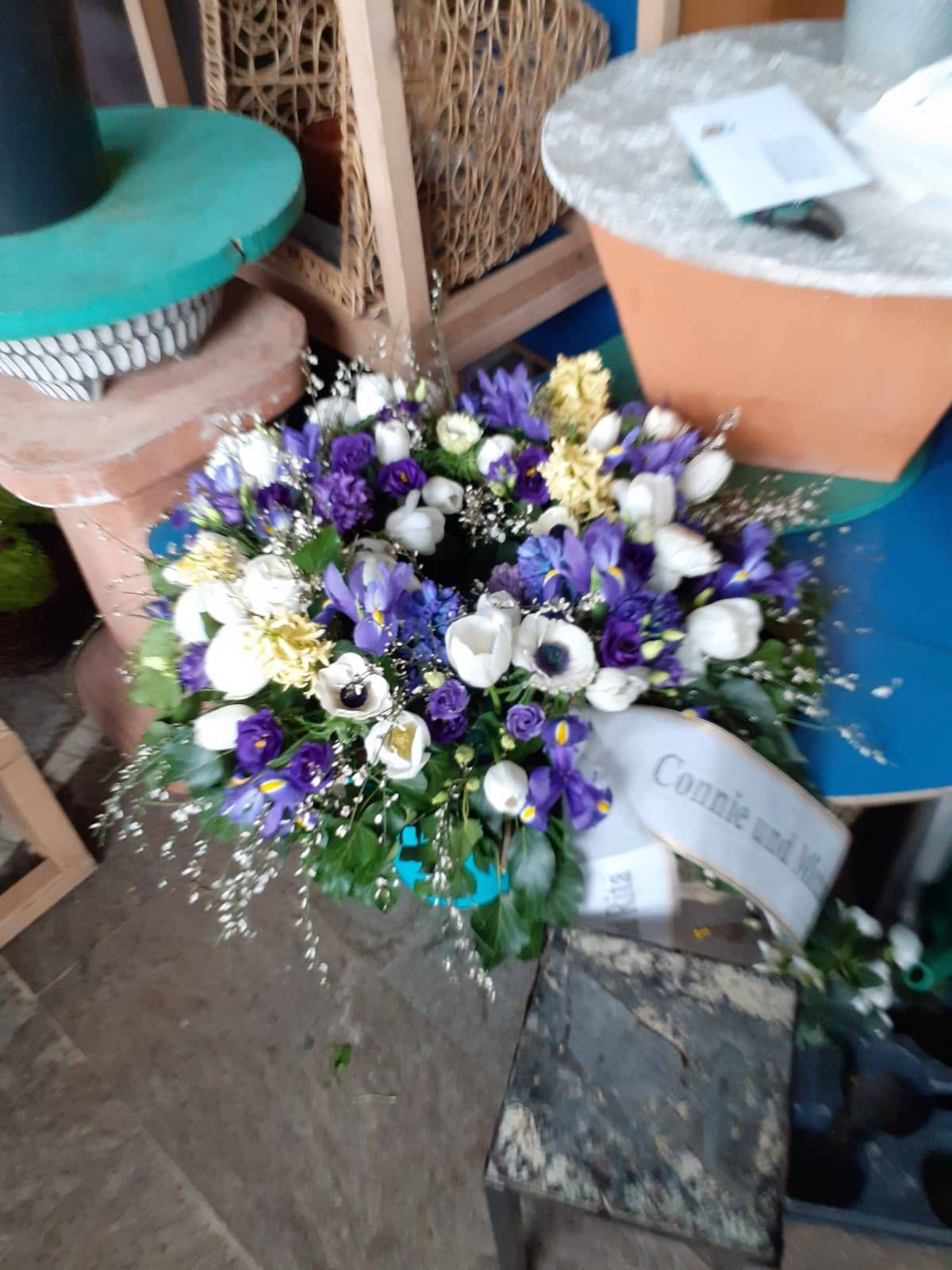 Blumenkranz mit lila, weißen und gelben Blumen, Schleife mit Text.