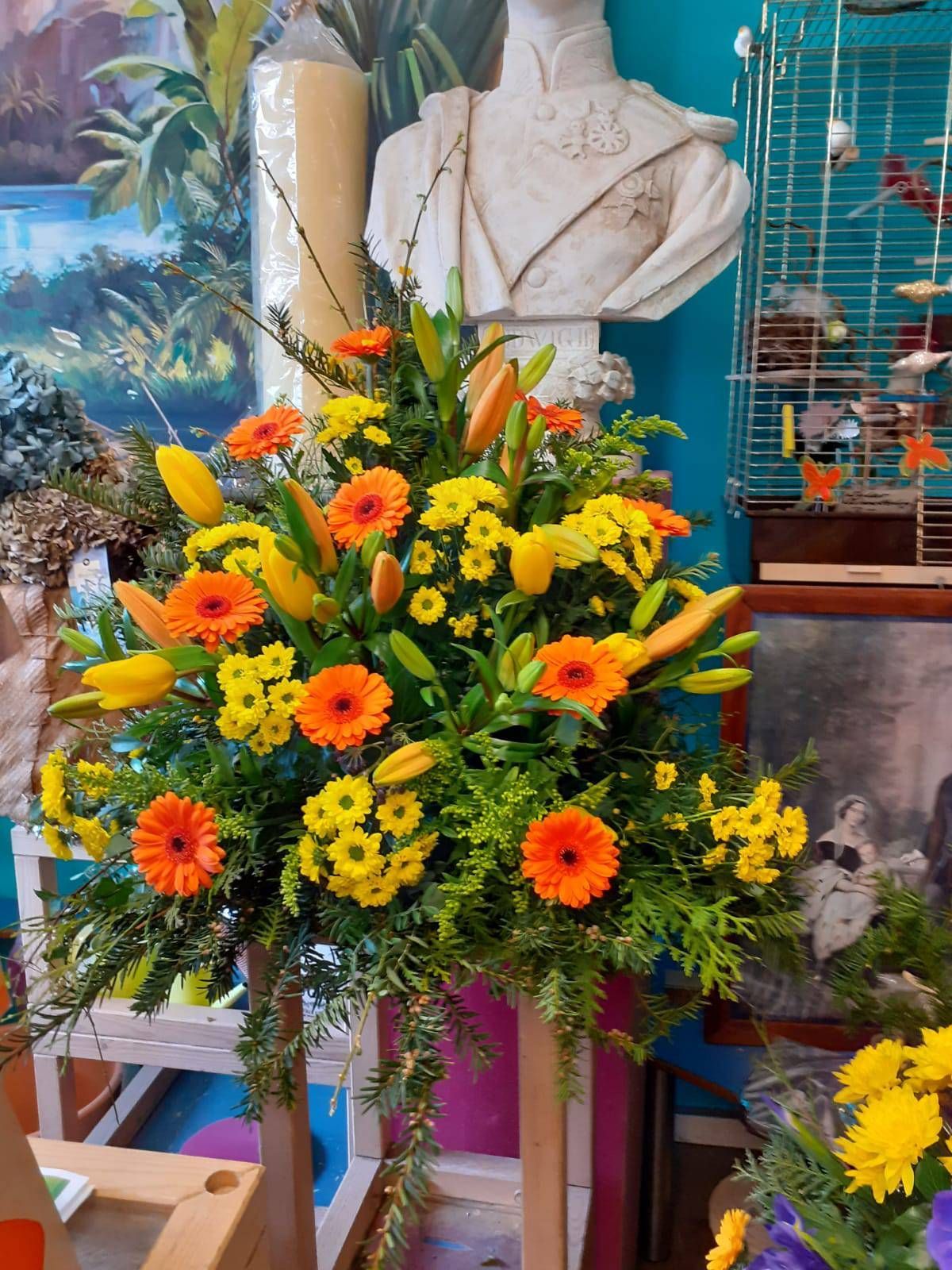 Blumenarrangement mit orangefarbenen Gerbera-Gänseblümchen, gelben Blüten und Lilien auf einem Holzständer.