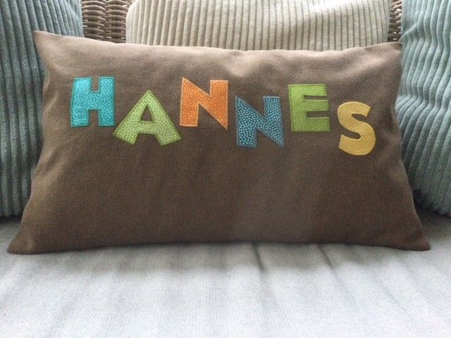 Braunes Kissen mit bunten „HANES“-Buchstaben auf einem gestreiften Sofa