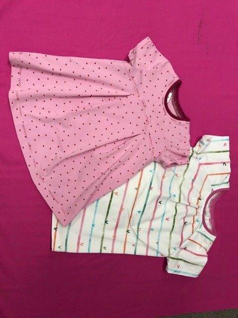 Zwei Babykleider, ein rosa gepunktetes und ein weiß gestreiftes, liegen auf einem rosa Hintergrund.