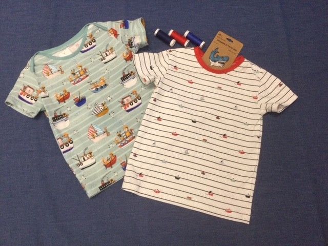 Zwei Kinder-T-Shirts auf blauem Grund, eines hellblau mit Booten und eines weiß mit roten Streifen, jeweils mit einem angebrachten Etikett.