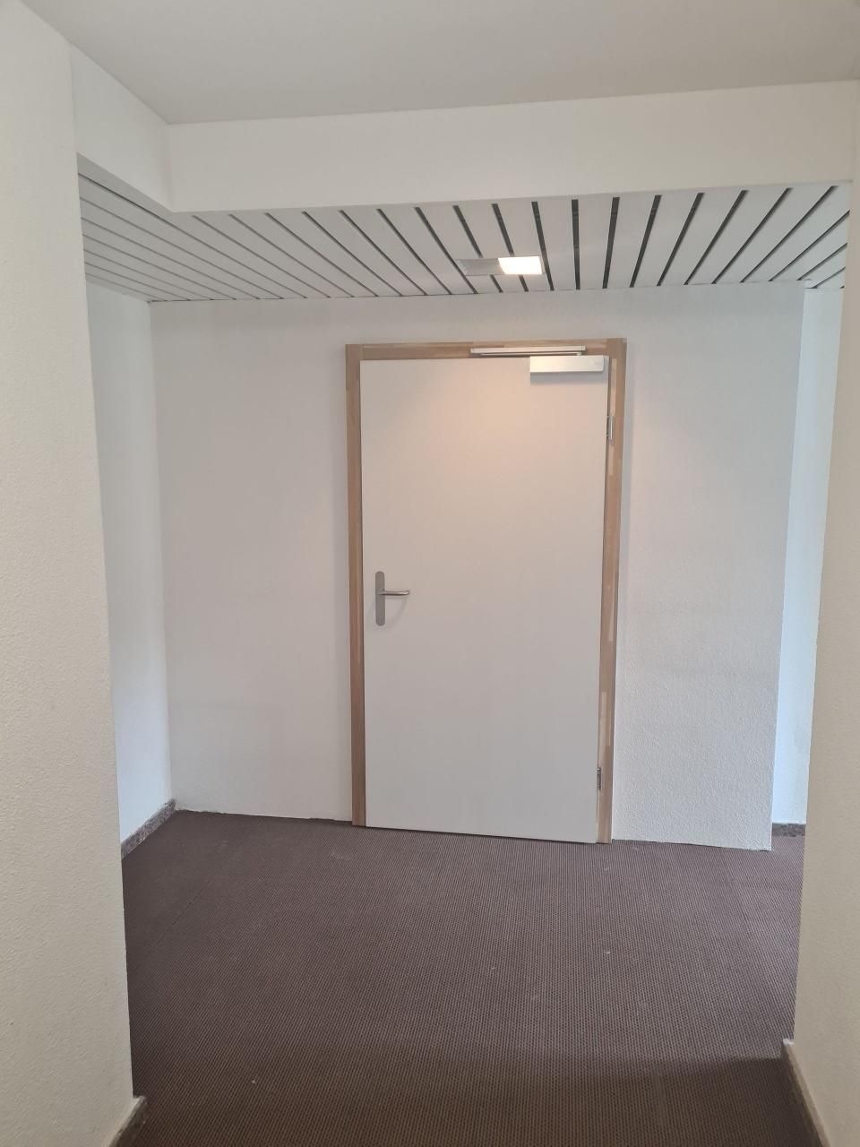 Leerer Büroflur mit geschlossener weisser Tür, grauem Teppichboden und gestreifter Deckenverkleidung.