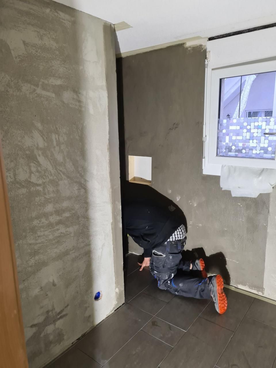 Ein Arbeiter kniet in einem grauen, unfertigen Raum in der Nähe eines Fensters und montiert oder überprüft eine Wandarmatur.