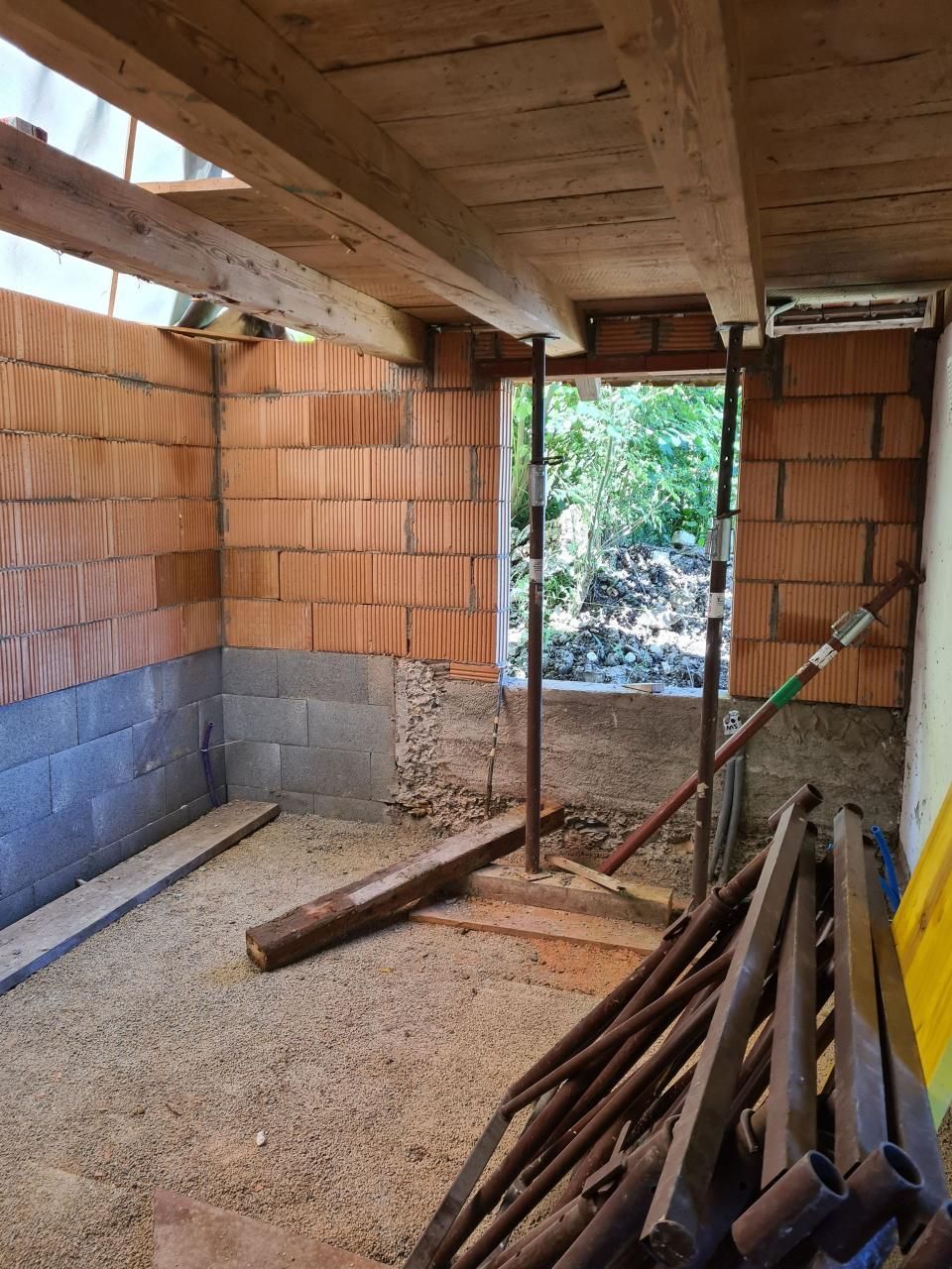Innenansicht eines unfertigen Backsteinraums mit freiliegenden Holzbalken, Kiesboden und gestapelten Rohren in der Nähe einer Tür.
