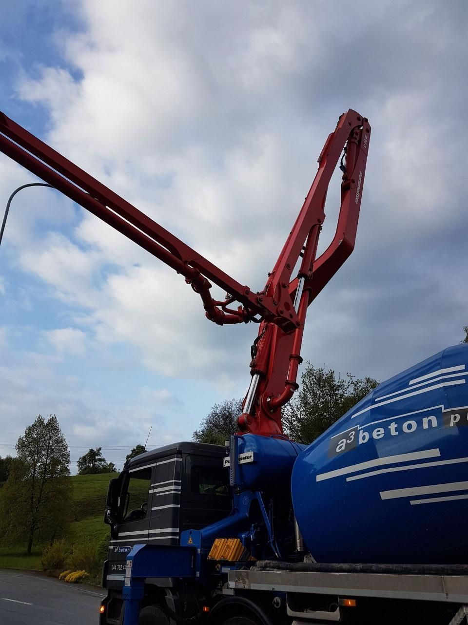 Ein roter Betonpumpenwagen mit ausgefahrenem Ausleger vor bewölktem Himmel am Strassenrand.