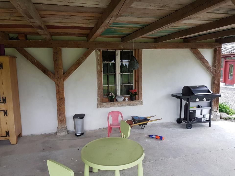 Überdachte Terrasse mit grünem Tisch, rosa Stuhl, Grill und hölzernen Stützbalken