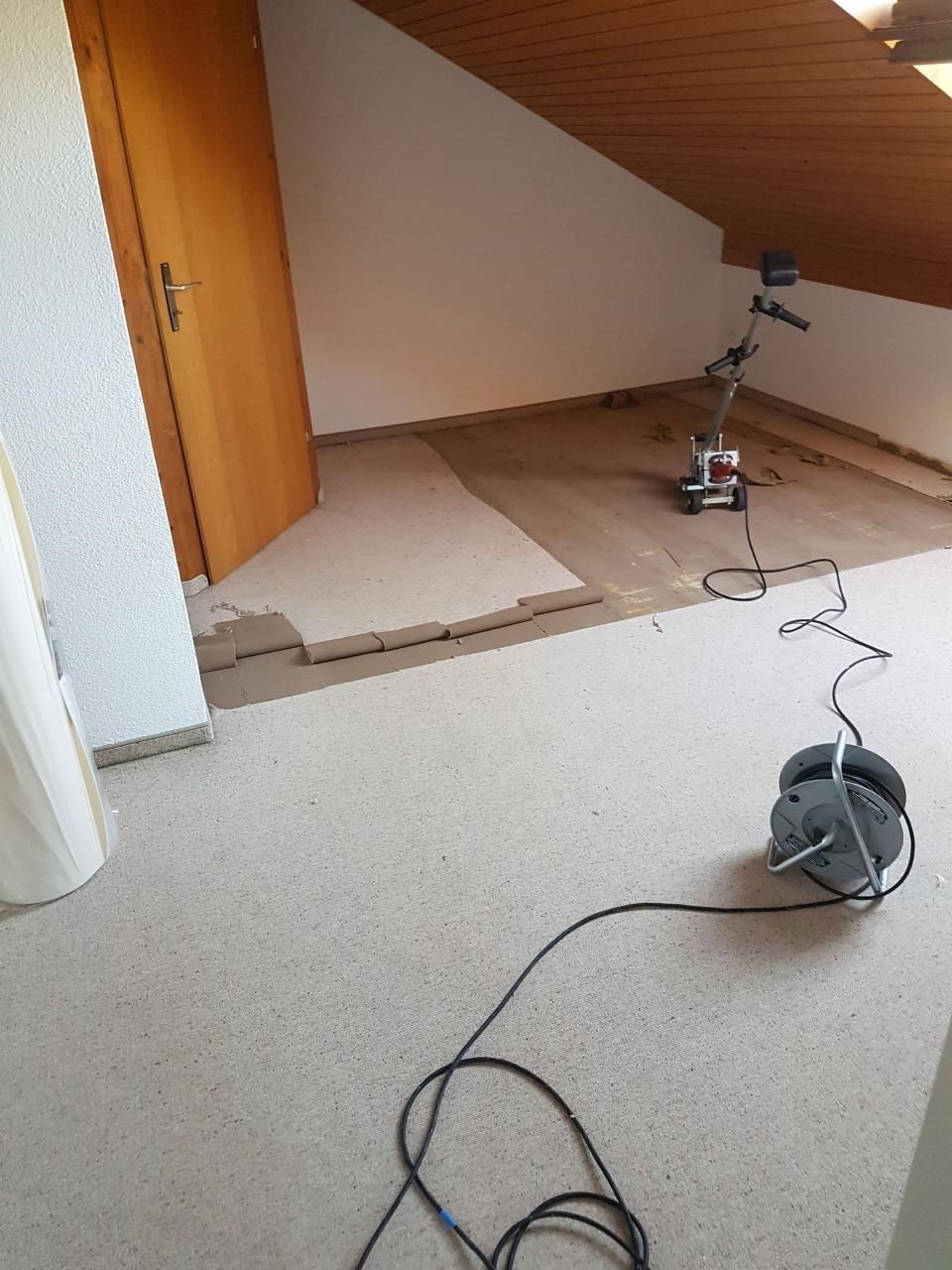 Zimmer mit beschädigtem Fussboden im Türbereich, der freiliegende Unterboden und ein kabelgebundenes Bodenreinigungsgerät zeigt.