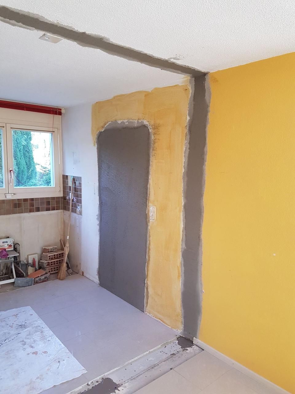 Teilweise renoviertes Zimmer mit gelben und grauen Wänden, einer Tür und einem Fenster, durch das Licht hereinfällt.