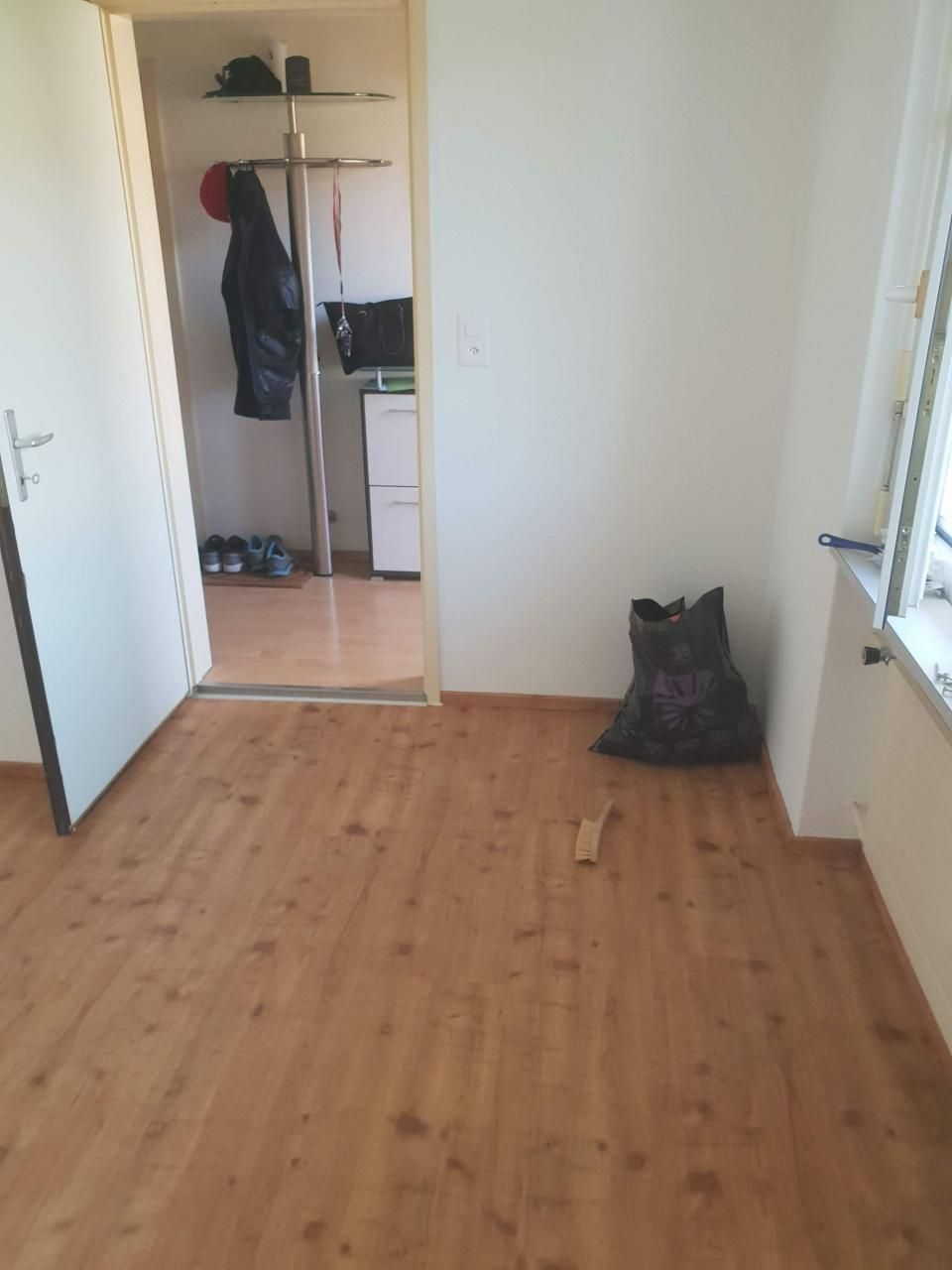 Leeres Zimmer mit Holzboden, offener Tür, Garderobe und einer schwarzen Tasche an der Wand