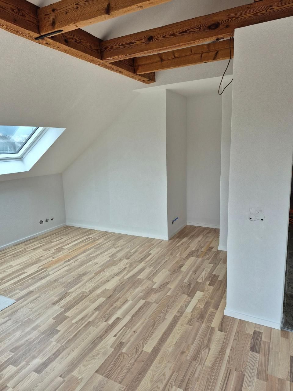Leeres Dachgeschosszimmer mit Dachfenster, freiliegenden Holzbalken und hellem Holzboden