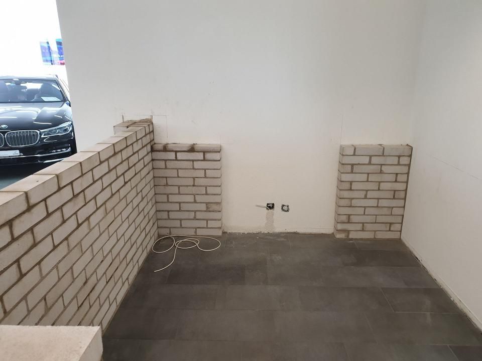 Leerer Raum mit weissen Wänden, dunklem Fliesenboden und niedrigen, halbhohen Ziegelwänden an der linken und hinteren Seite.
