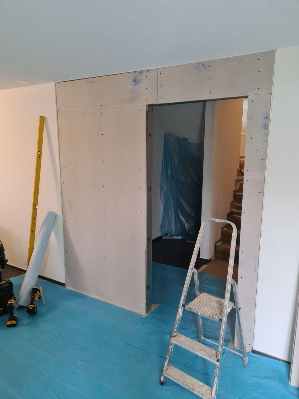 Teilweise fertiggestellte Innenwand mit Türöffnung, blauem Boden, Leiter und Werkzeugen auf einer Baustelle