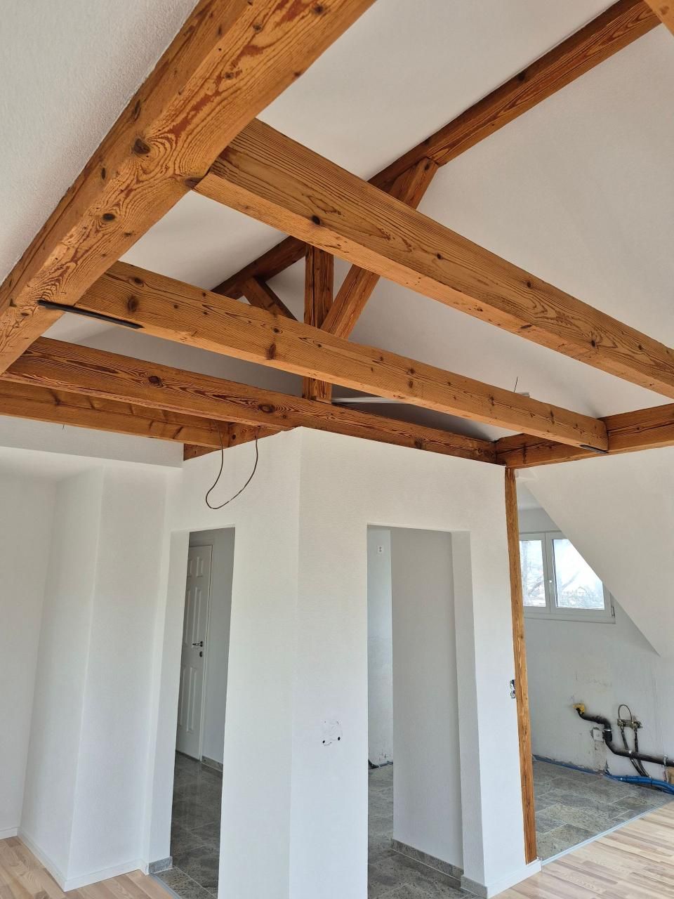 Weisser Innenraum unter freiliegenden Holzbalken, mit unverputzten Wänden und einem Durchgang zu einem Küchenbereich.