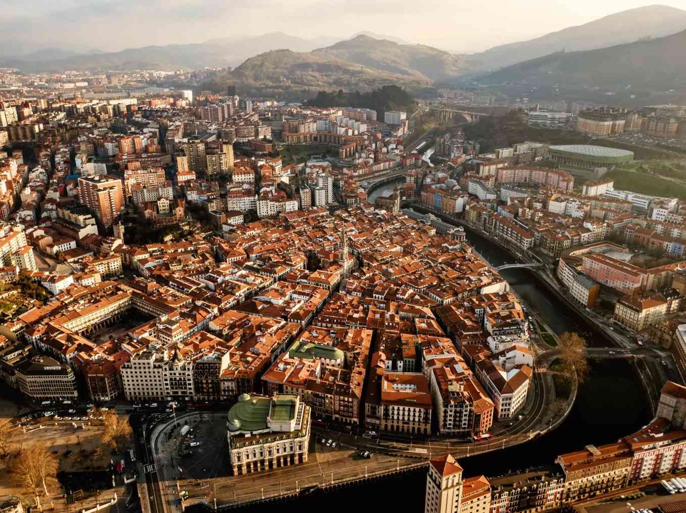 Vista aérea de una ciudad densamente poblada con edificios de tejados rojos, río, puente y montañas en la distancia.