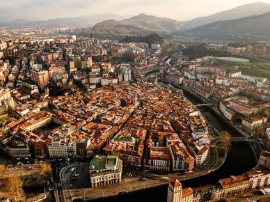 Vista aérea de una ciudad densamente poblada con edificios de tejados rojos, río, puente y montañas en la distancia.