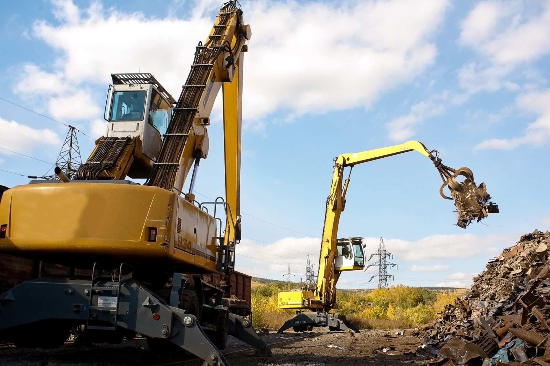 Dos excavadoras levantando chatarra en un desguace bajo un cielo azul.