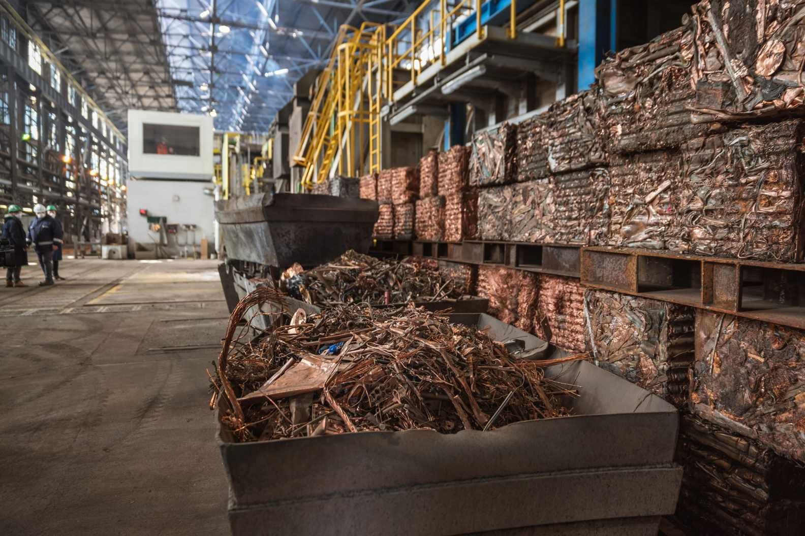 Almacén industrial con fardos de chatarra de cobre apilados y una gran pila de chatarra metálica en primer plano.