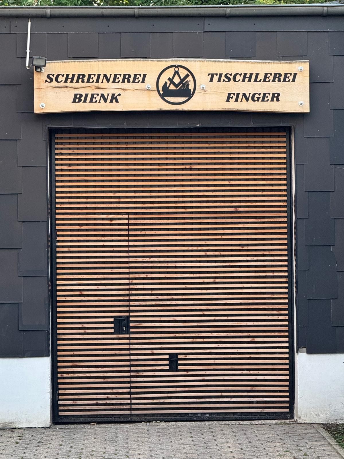 Schild über einer hölzernen Werkstatttür; darauf steht „Schreinerei Bienk Tischlerei Finger“ mit einem Logo