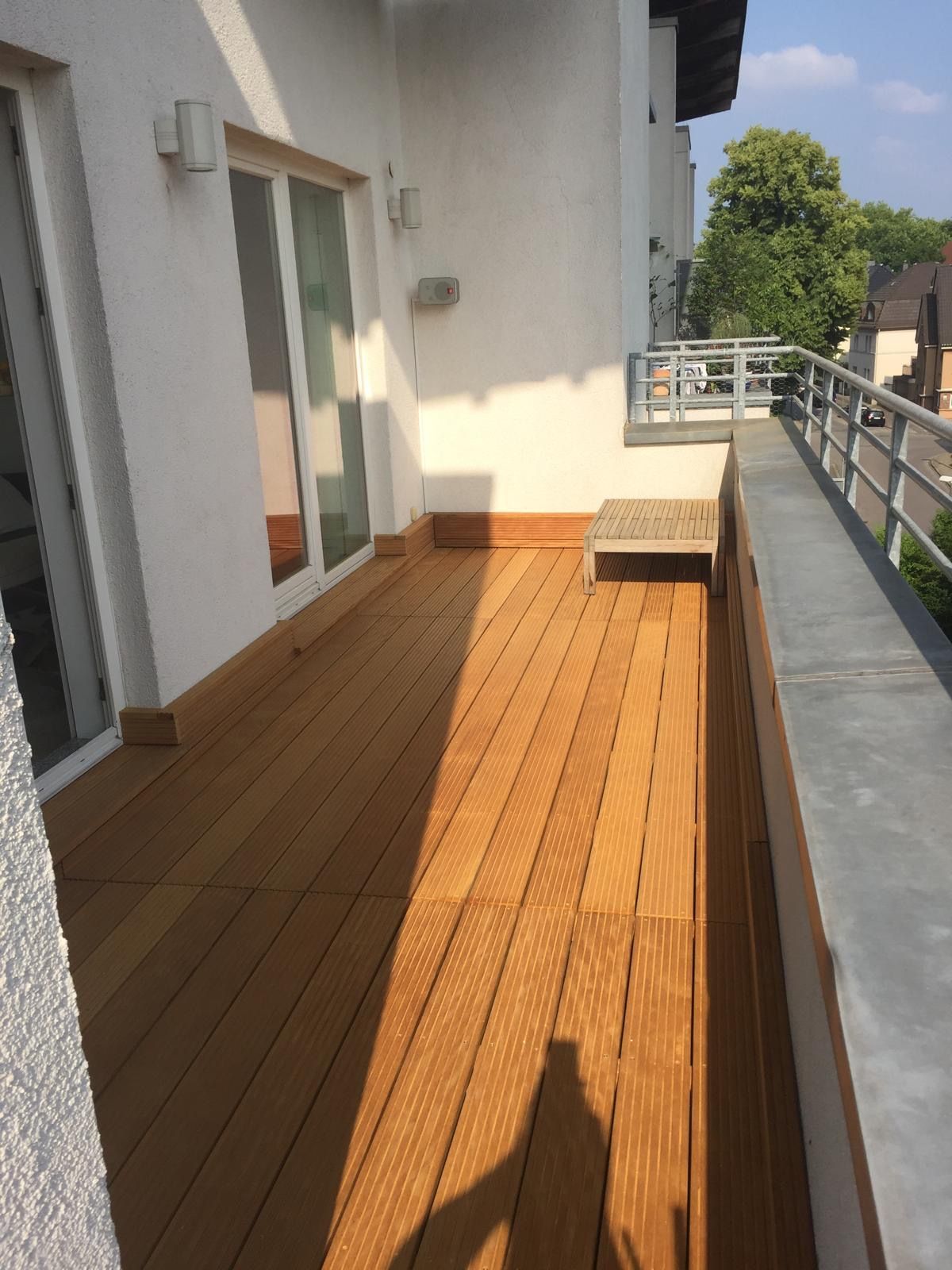 Holzterrasse mit Geländer, weißer Wand, Glastüren und kleiner Bank im Freien.