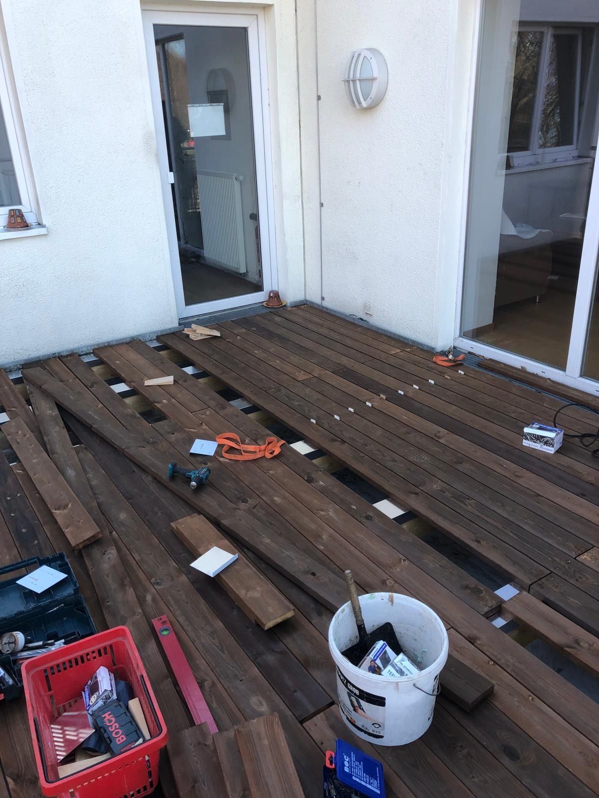 Terrassenbau: Holzdielen werden auf einer im Bau befindlichen Terrasse verlegt. Werkzeug und ein Eimer sind vorhanden.