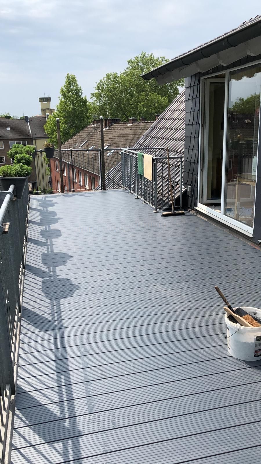Balkon mit grauem Metallboden, Geländer und Blick auf Dächer und Bäume.