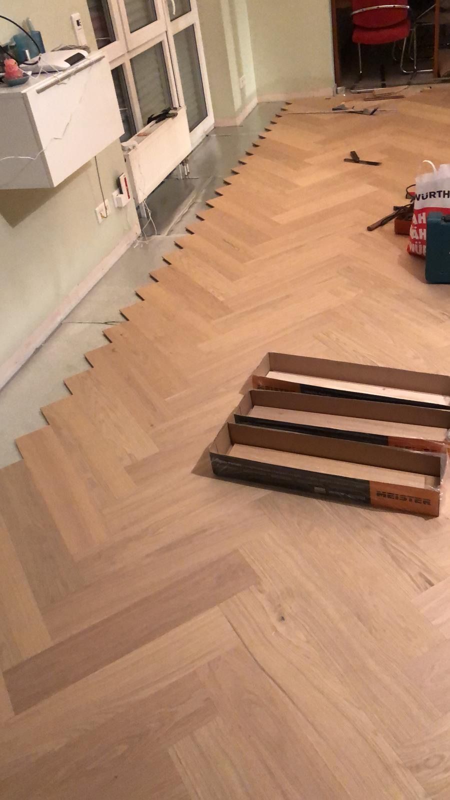 Die Verlegung eines Fischgrätparketts ist im Gange; Dielen, Werkzeuge und erste Montagearbeiten sind bereits erfolgt.