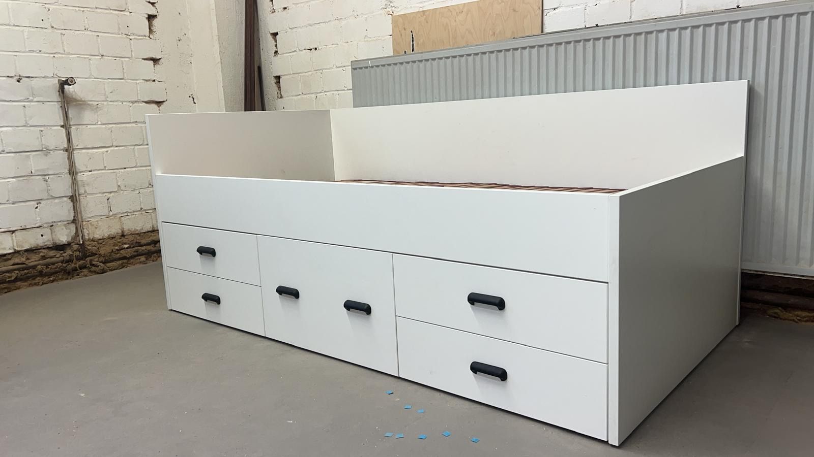 Weißes Bettgestell mit sechs Schubladen, vor einer Backsteinwand.