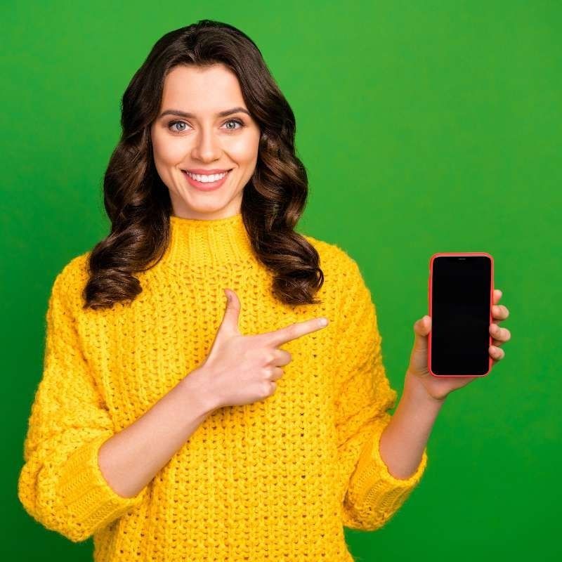 Una mujer con un suéter amarillo sostiene un teléfono celular y lo señala.
