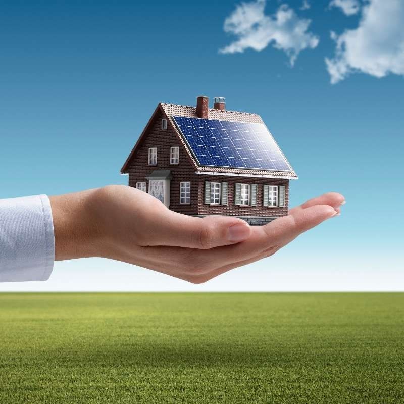 Una mano sostiene una pequeña casa con paneles solares en el techo.