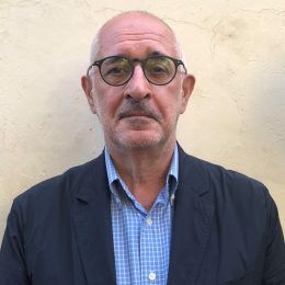 A bald man wearing glasses and a suit is standing in front of a wall.