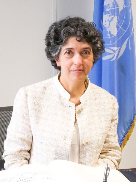 A woman in a white jacket is sitting in front of a blue flag.