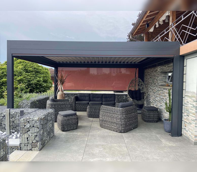 Terrasse extérieure avec mobilier gris sous une pergola sombre, un mur en pierre et un fauteuil suspendu.