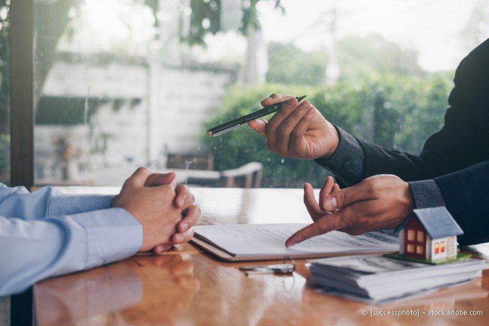 Zwei Personen sitzen an einem Tisch und sichten Dokumente. Eine Person zeigt mit einem Stift auf ein Blatt Papier; darauf ist ein kleines Hausmodell zu sehen.