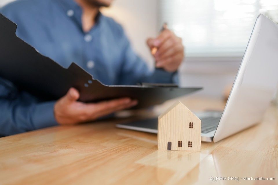 Ein Mann sichtet Dokumente, ein kleines Hausmodell und einen Laptop auf einem Holzschreibtisch.
