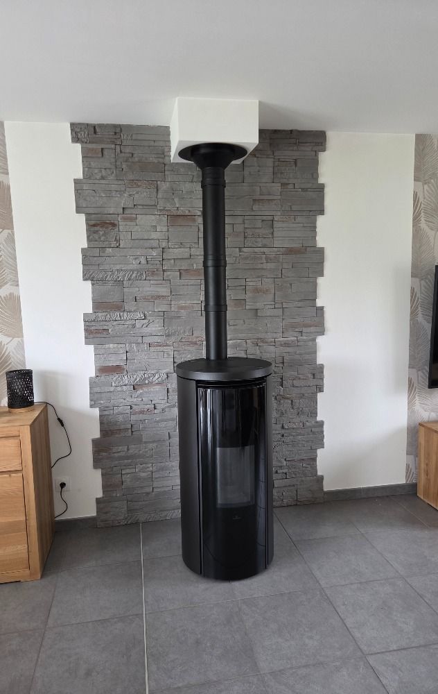 Poêle à bois noir brûlant dans un coin beige d'une pièce, avec conduit de cheminée et socle noirs.