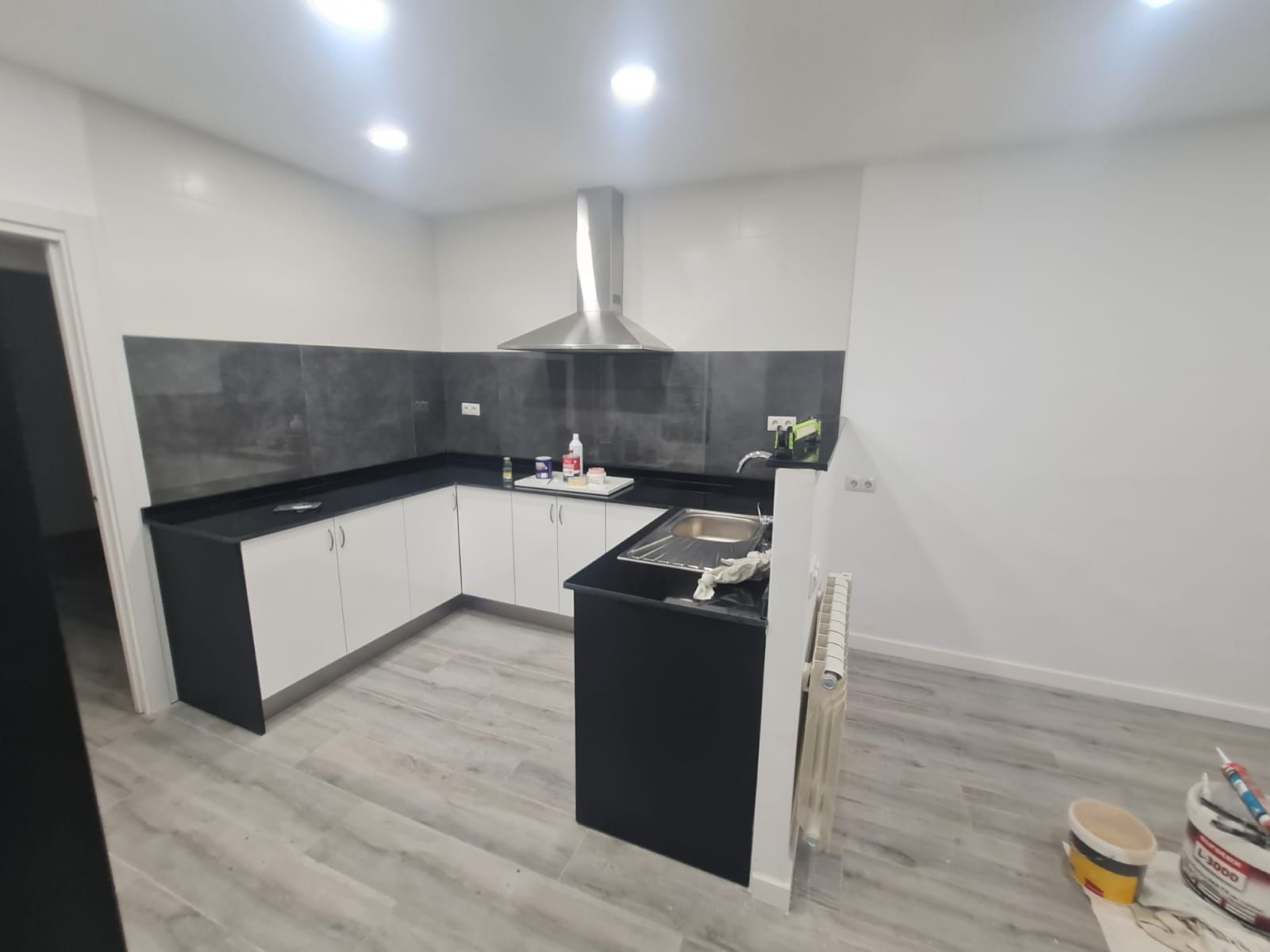 Cocina moderna en forma de L con gabinetes blancos y negros, encimera negra y campana extractora de acero inoxidable.