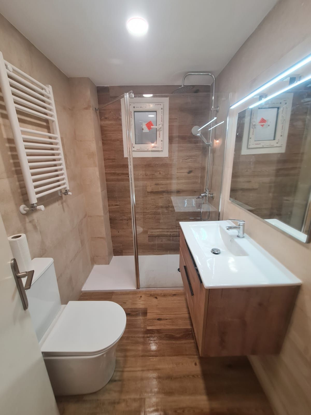Pequeño baño con suelo y paredes imitación madera, ducha, lavabo e inodoro; tonos blanco y marrón.