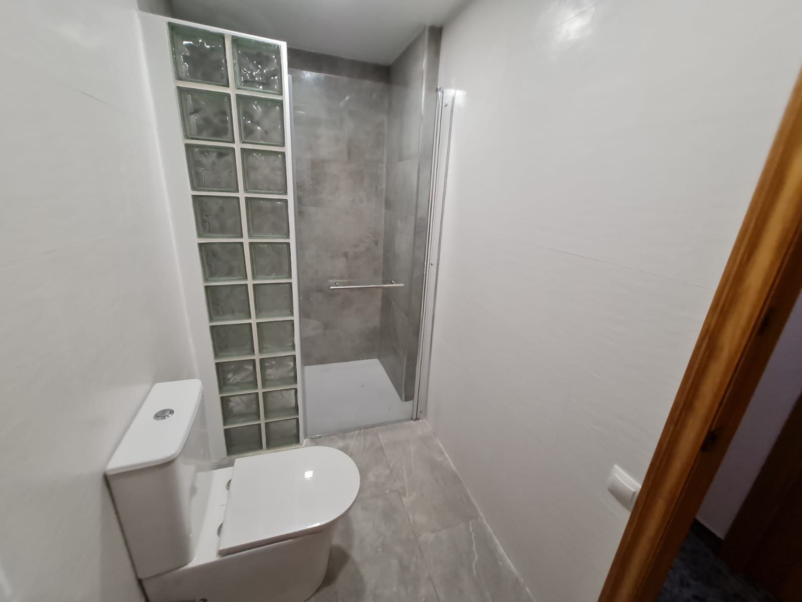 Baño con ducha de azulejos grises, pared de bloques de vidrio, inodoro blanco y piso de baldosas grises.