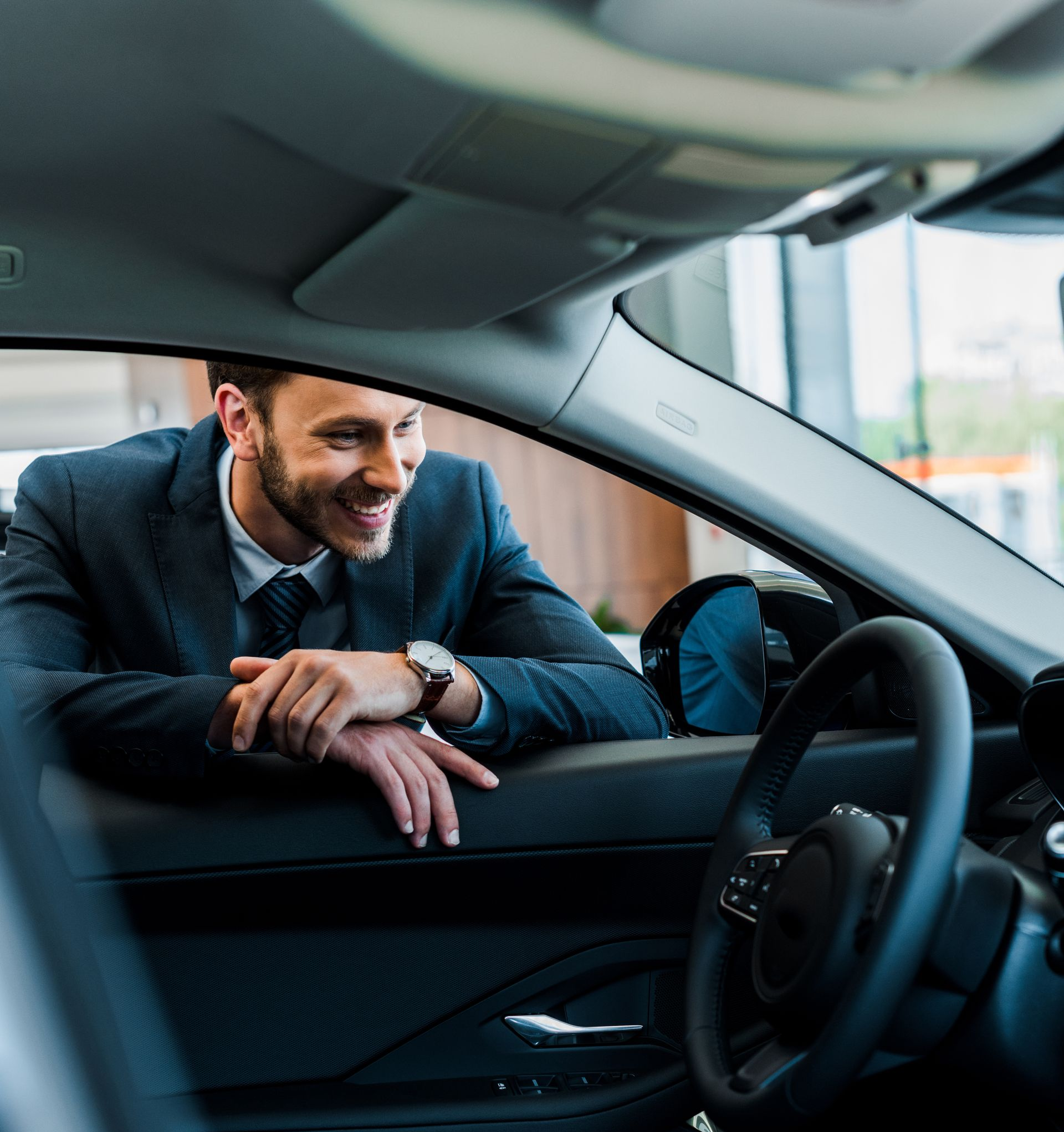Un homme en costume sourit, penché dans une voiture, les mains sur le cadre de la portière, dans une salle d'exposition.