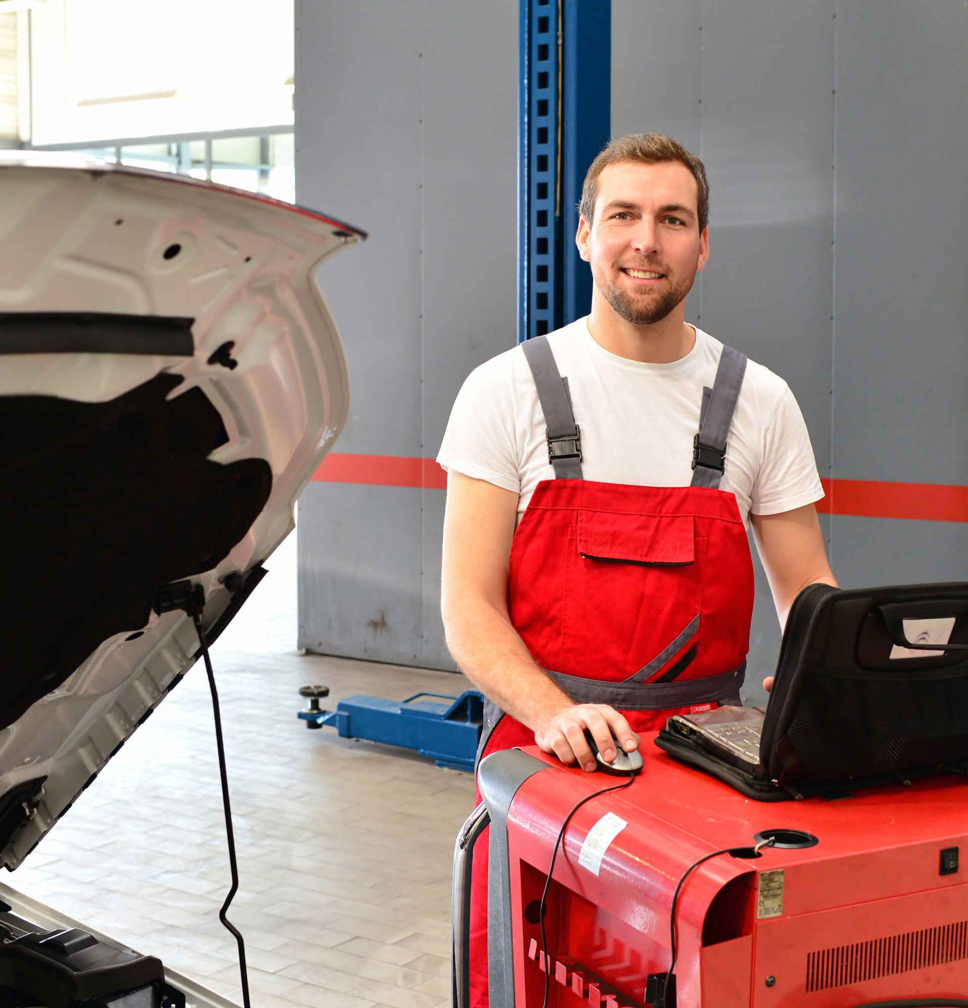 Un mécanicien en salopette, souriant, utilise un ordinateur portable dans un garage, le capot d'une voiture ouvert.