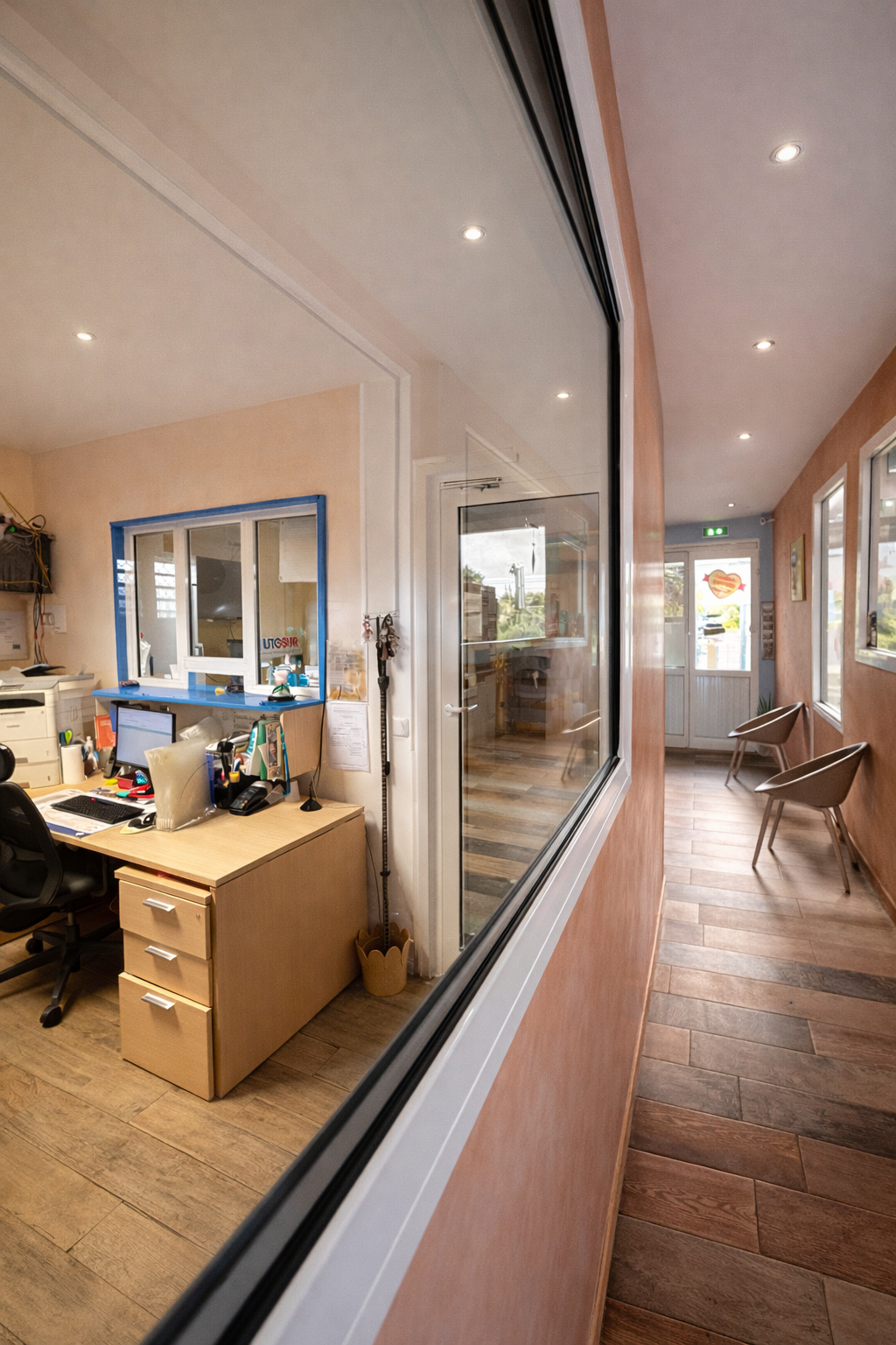 Vue à travers une cloison vitrée donnant sur un bureau avec un bureau et un ordinateur, le long d'un couloir avec des chaises et un sol carrelé.