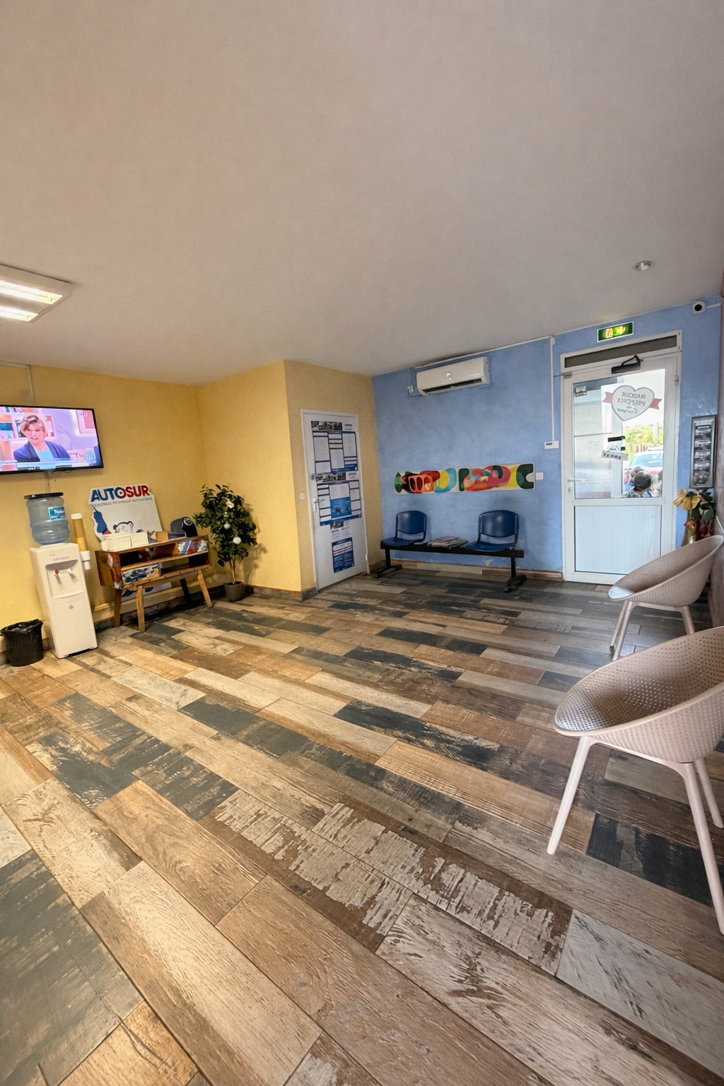Une salle d'attente avec des chaises claires, une télévision, une fontaine à eau et un sol imitation bois.
