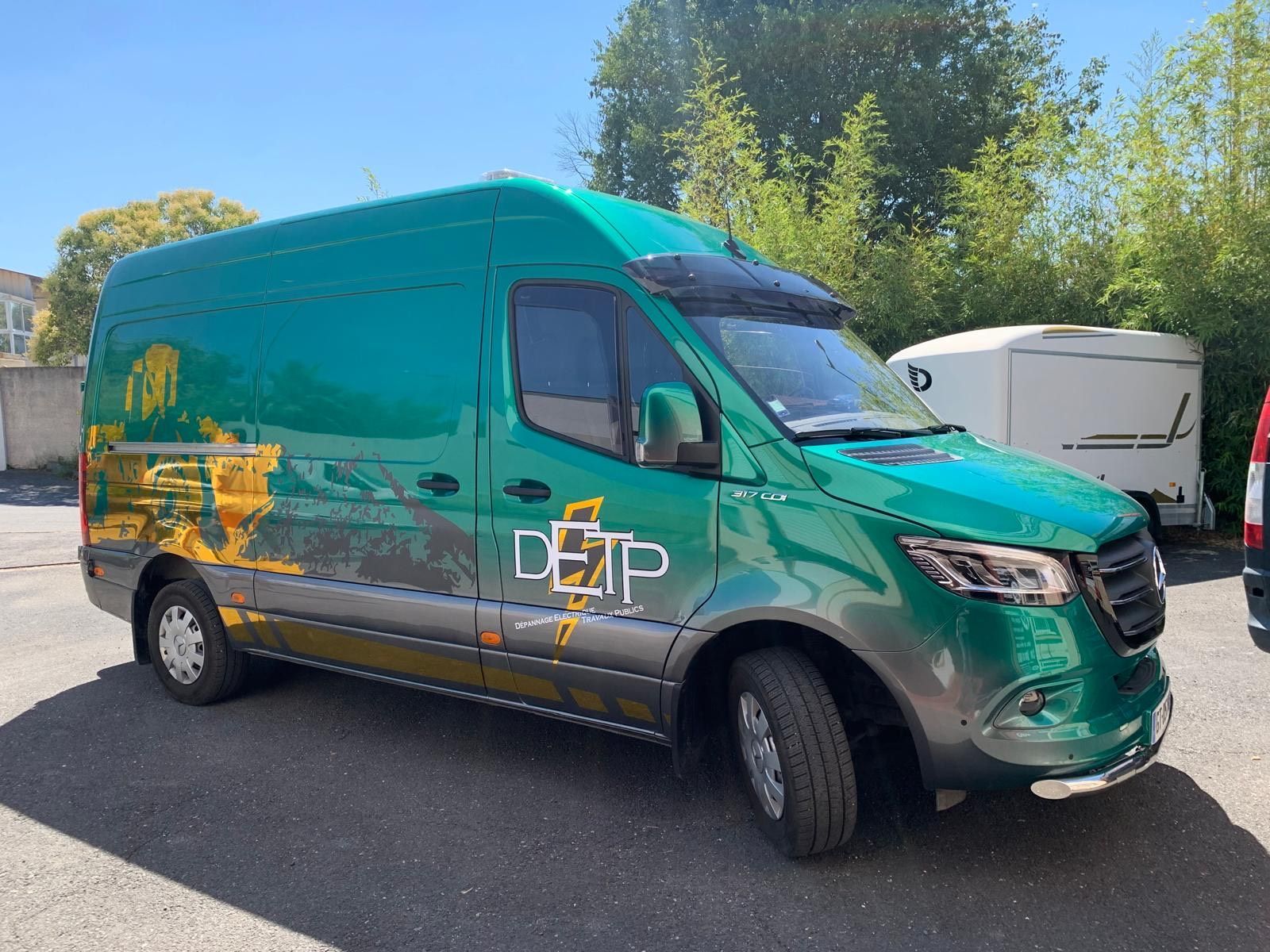 Une camionnette de livraison verte est garée à l'extérieur en plein soleil, avec des arbres et une remorque blanche en arrière-plan.