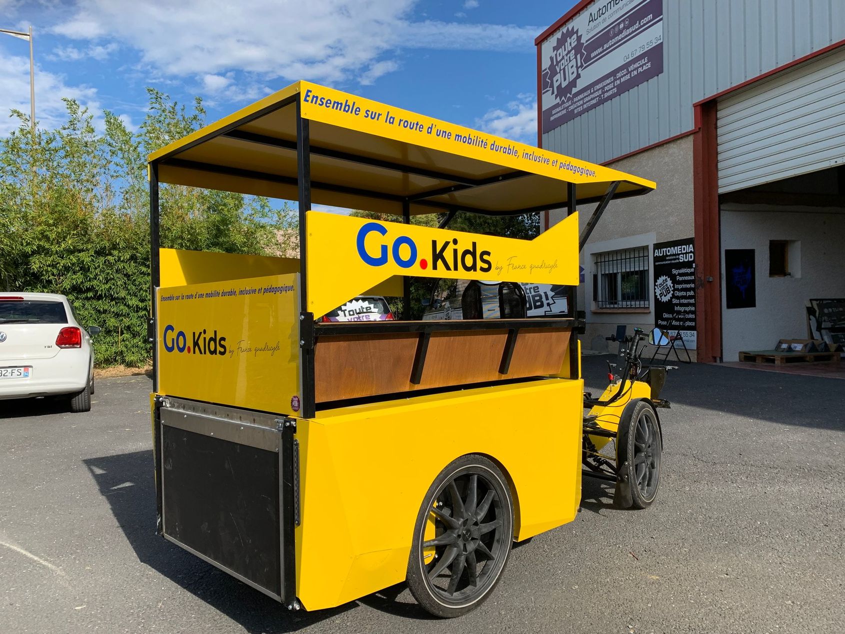 Chariot publicitaire jaune GoKids stationné devant un bâtiment sur un terrain ensoleillé.