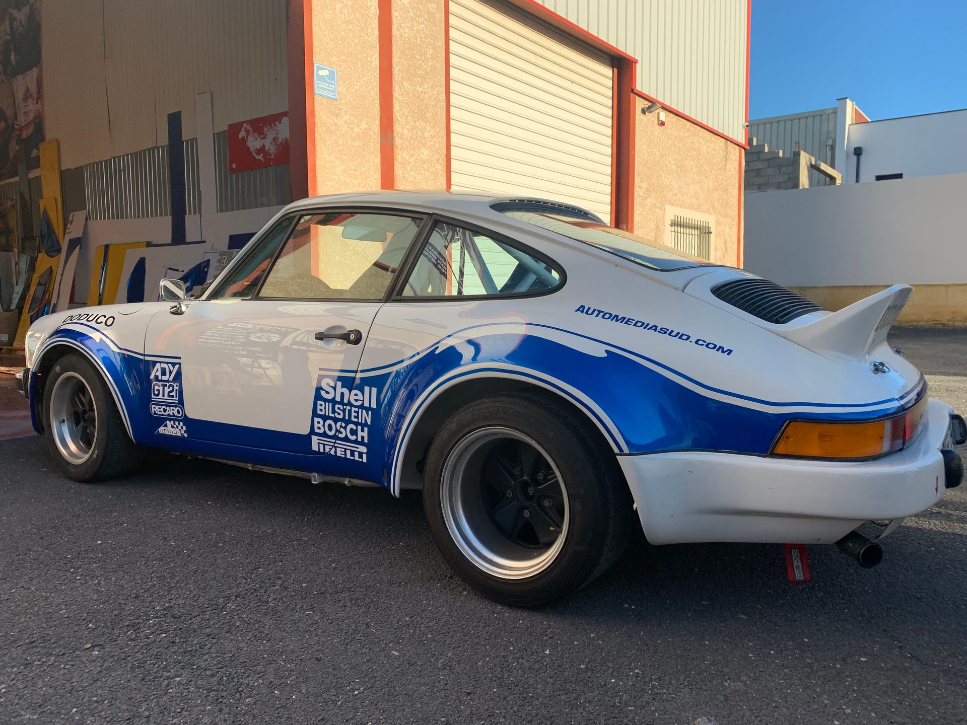 Porsche 911 blanche et bleue garée devant un bâtiment, vue de l'arrière.