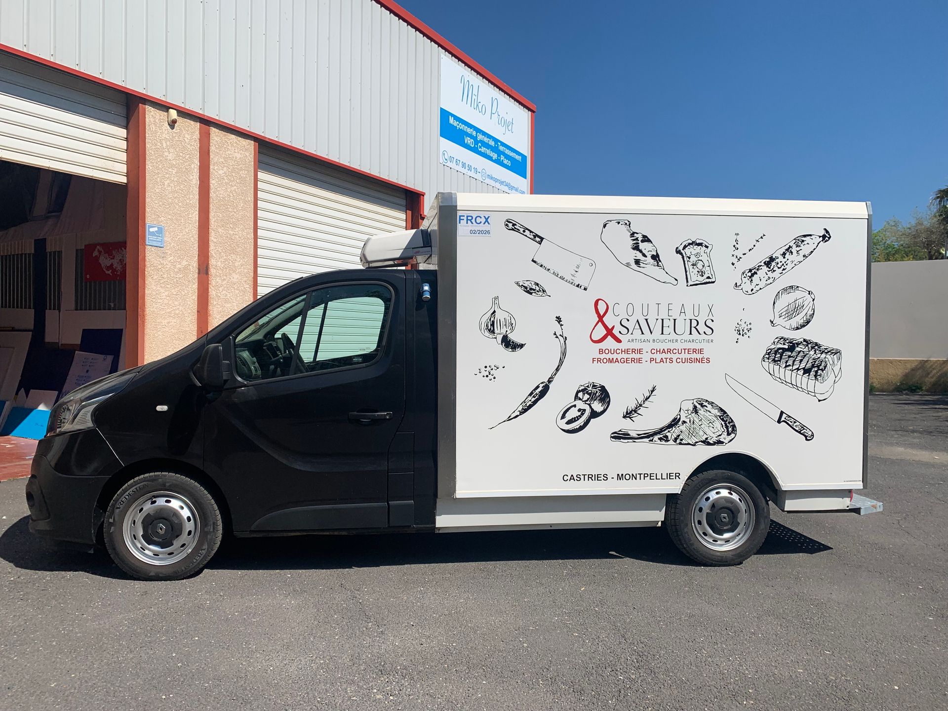 Une camionnette de livraison noire avec une caisse de chargement blanche est garée devant un entrepôt.