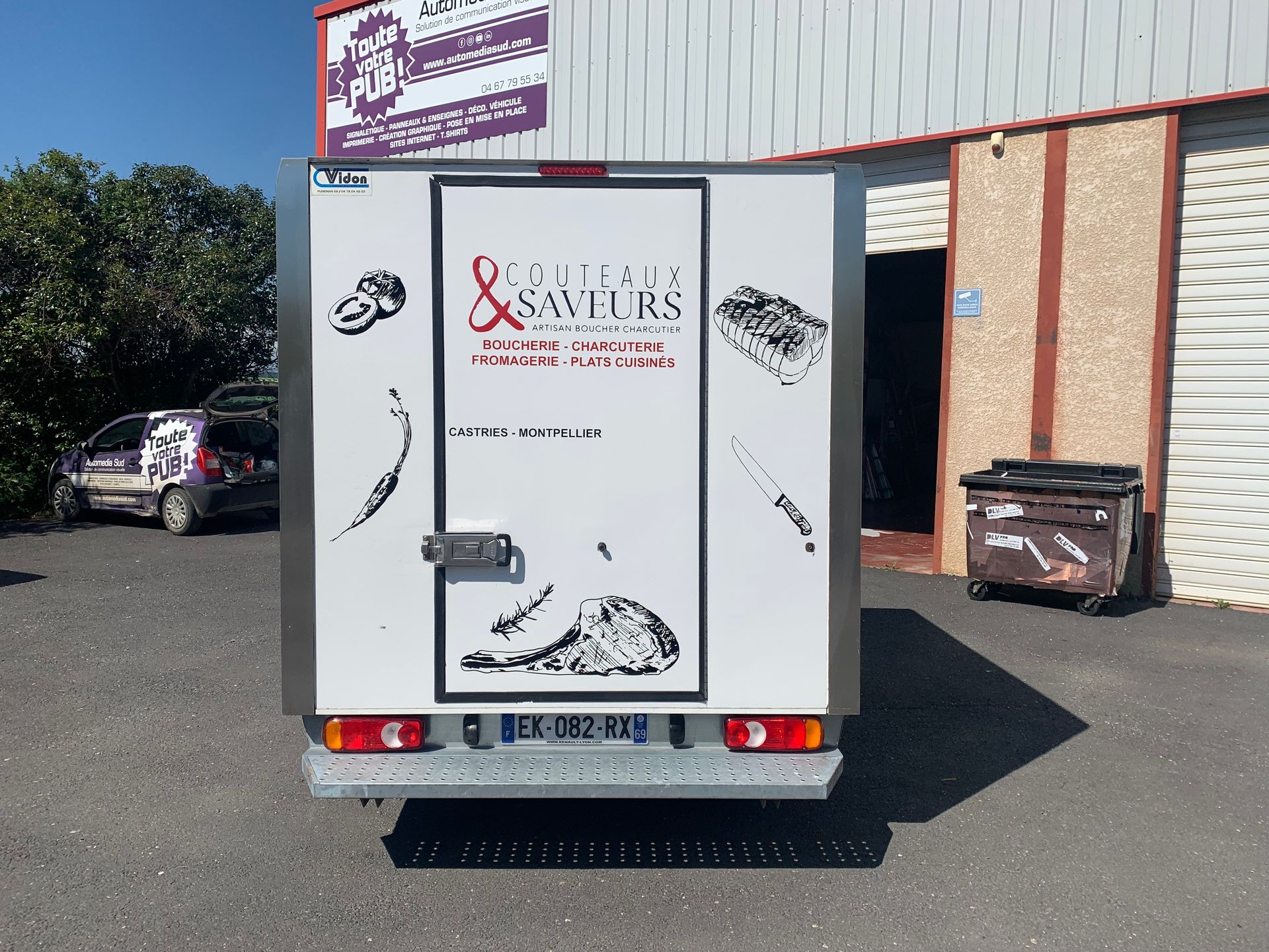 Camionnette de livraison blanche stationnée près d'un bâtiment, vue arrière avec logo et graphismes de l'entreprise.