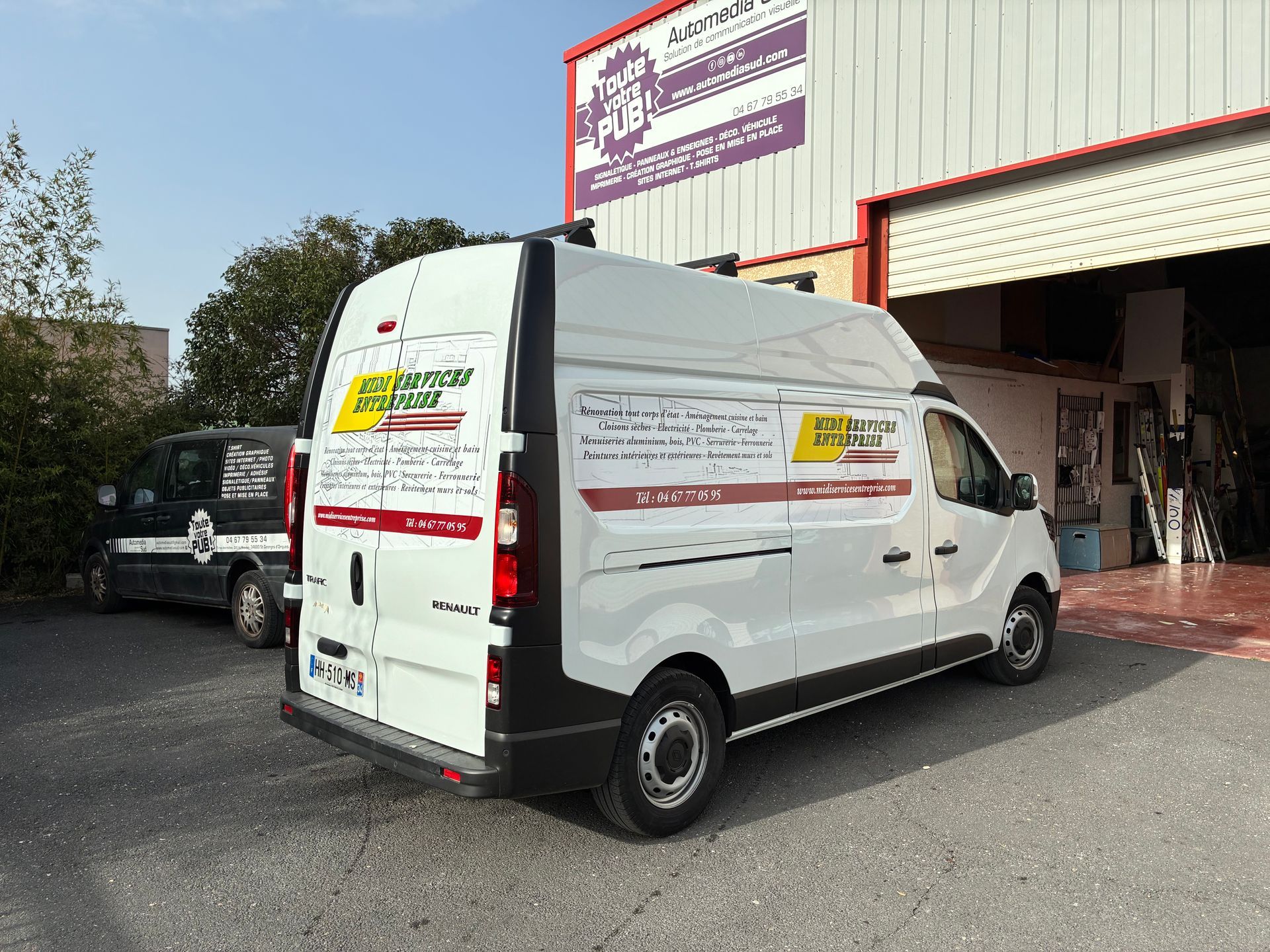 Une camionnette de livraison blanche est garée devant l'entrée d'un immeuble avec des panneaux, et une voiture noire se trouve à proximité.