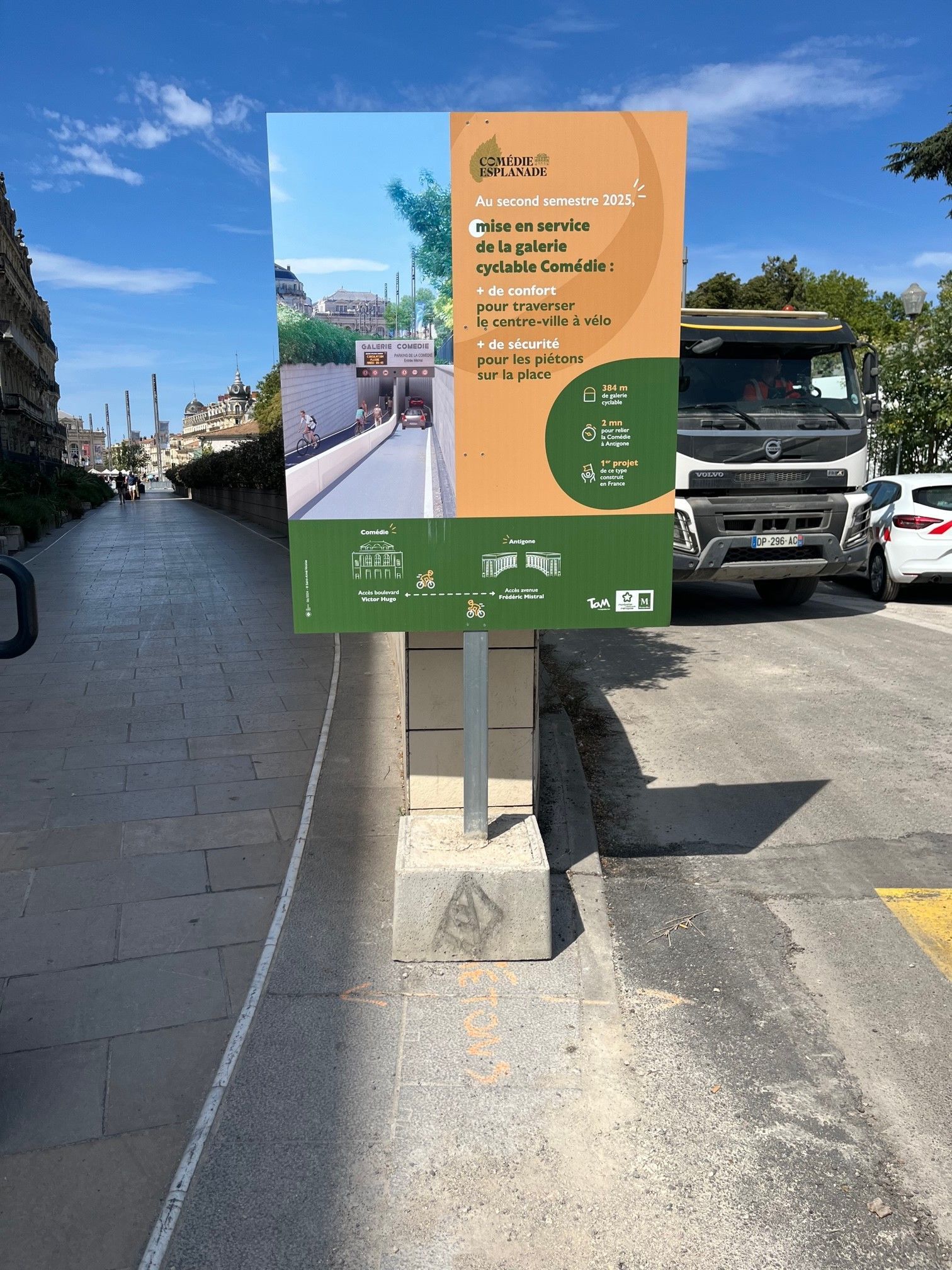Panneau d'affichage extérieur pour un événement, avec un design orange et vert, sur un trottoir au bord d'une route sous un ciel bleu.