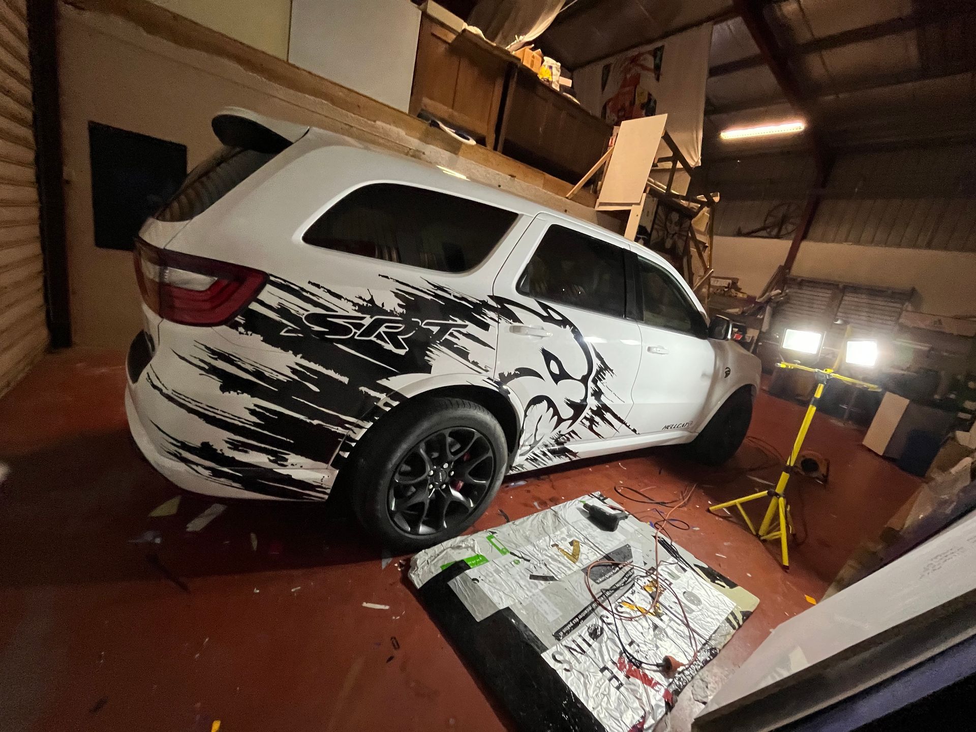 Un SUV blanc orné d'un dessin de peinture noire est garé dans un garage ; des journaux jonchent le sol et une lampe de chantier est allumée à proximité.